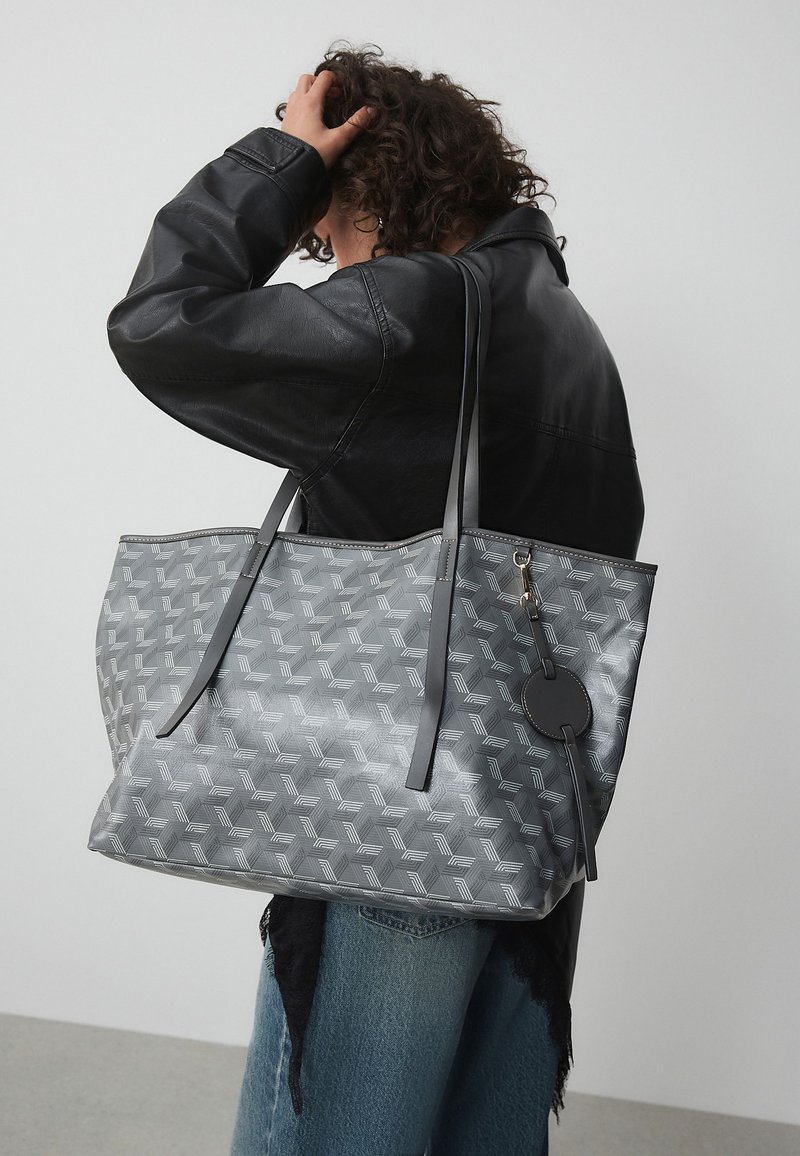 Personne aux cheveux bouclés portant une veste en cuir noire et tenant un grand sac cabas gris à motifs avec de longues anses sur l'épaule.