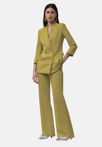 Completo giacca doppiopetto e pantaloni a gamba larga di colore giallo senape. La giacca ha un colletto a revers e tasche frontali; il modello indossa scarpe argentate.