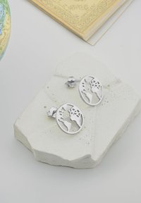 Silvriga örhängen formade som jorden, med utskurna kontinenter, som vilar på en texturerad vit stenbas.