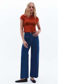 Červený krajkový top s krátkým rukávem zkombinovaný s modrými džíny s vysokým pasem a širokými nohavicemi, na bílém pozadí.