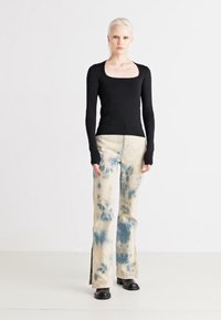 Svart langermet topp med firkantet utringning, kombinert med blå tie-dye flared jeans og svarte ankelstøvler. Enfarget bakgrunn, helkroppsbilde.