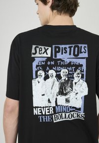 T-shirt en coton noir avec un graphisme représentant quatre personnages en blanc et bleu, accompagné du texte "Sex Pistols" et "NEVER MIND THE BOLLOCKS", sérigraphié.
