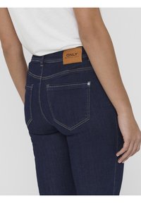 Persona con vaqueros de mezclilla azul oscuro que tienen bolsillos traseros y una etiqueta de marca de cuero que dice "ONLY" cerca de la cinturilla.