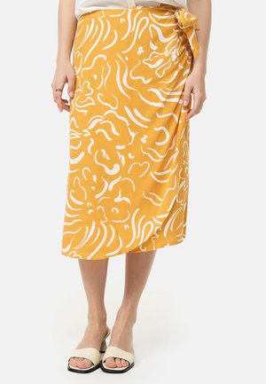 Vrouw met een gele wikkelrok met witte abstracte patronen, een strik aan de zijkant, witte sandalen en een witte top, staand tegen een effen achtergrond.