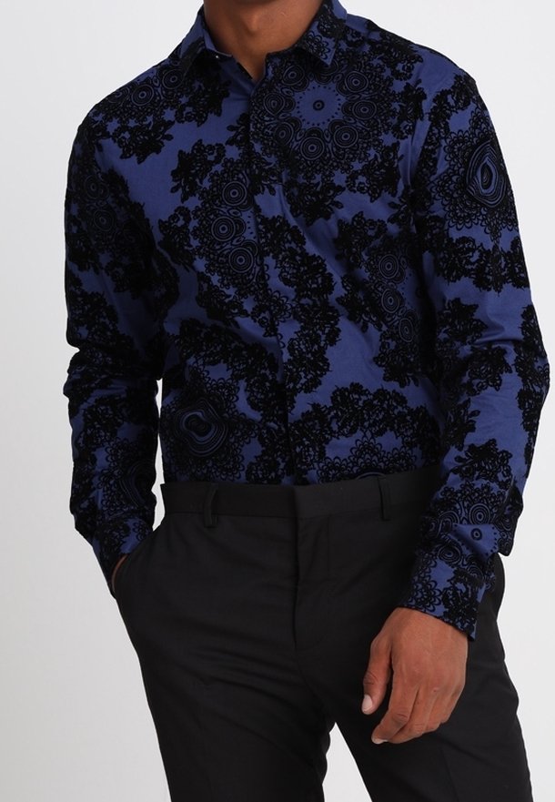 Homme portant une chemise bleu marine à manches longues avec des motifs floraux noirs complexes, associée à un pantalon noir, debout avec une main dans la poche.