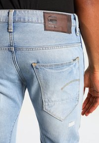 Ljust blå denimjeans med en brun läderetikett, med en bakficka med sömdetaljer och synlig slitage på tyget.