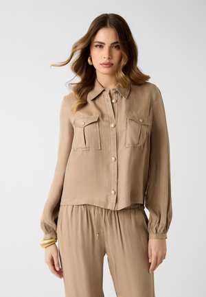 Femme aux cheveux ondulés portant une chemise beige à boutons avec deux poches poitrine et un pantalon ample assorti, accessoirisée avec des boucles d'oreilles et un bracelet en or.