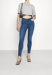Camiseta gris ajustada, jeans azules ceñidos y sandalias de tacón verdes. Incluye texturas suaves, un ajuste recto y accesorios sutiles.