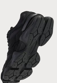 Chaussure de sport noire avec une tige texturée, une semelle rembourrée et une semelle extérieure en caoutchouc multidirectionnelle. Présente des détails de logo proéminents.