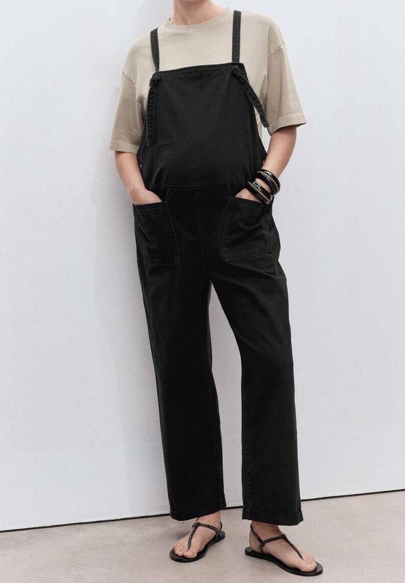 Personne portant une chemise beige à manches courtes, une salopette noire à jambes larges avec des poches, des bracelets noirs et des sandales noires, debout contre un mur blanc.