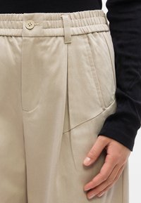 Μπεζ πλισέ παντελόνι με ελαστική μέση και κουμπί, συνδυασμένο με μαύρο μακρυμάνικο τοπ, το χέρι ακουμπά κοντά στην τσέπη.