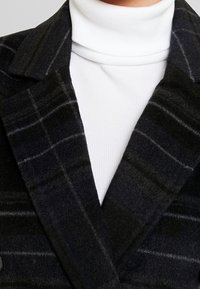 Czarny wełniany płaszcz w kratę z kołnierzem w klapach, z miękką fakturą i szarymi paskami; noszony na białym, prążkowanym golfie.