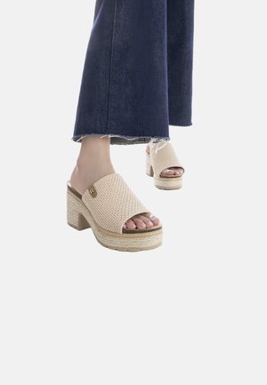 Sandali scivolosi beige intrecciati con una fascia larga, tacco spesso e dettagli in juta lungo la suola. Abbinati a pantaloni flare in denim scuro.