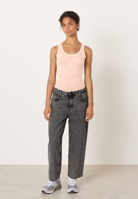 Jauna sieviete stāv, valkājot gaiši rozā bezroku topu, brīvas pelēkas džinsas un pelēkas un baltas sporta kurpes pie vienkāršas gaišas sienas.