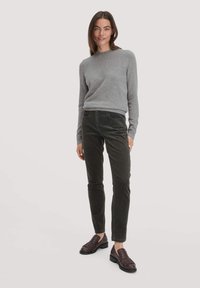 Vrouw die staat in een grijze lange mouwen trui, donkere olijfgroene corduroy broek en donkerbruine franjesloafers op een effen lichte achtergrond.