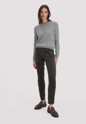 Frau steht mit einem grauen Langarm-Pullover, dunkeloliven Cordhose und dunkelbraunen Fransen-Loafers vor einem schlichten hellen Hintergrund.
