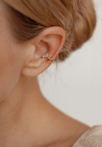 Silberne Creolenohrringe mit blattförmigen Akzenten, die zwei klare Edelsteine enthalten. Der Ohrring ist im Close-up an einem Ohr gestylt.