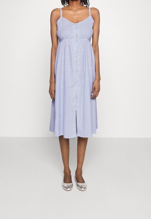 Vrouw met een mouwloze blauw-wit gestreepte midi-jurk met knopen en zilverkleurige ballerina's, staand tegen een witte achtergrond.