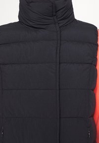 Svart pufferväst med hög krage, med quiltade sektioner. Dragkedjefickor på sidorna, i kombination med ett klart rött innerskikt.