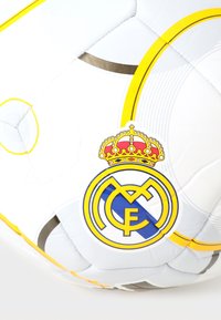 Balle de soccer blanche avec des accents jaunes et gris, arborant le logo du Real Madrid avec une couronne, texture lisse et panneaux traditionnels cousus.