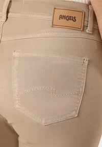 Primer plano de unos vaqueros de mezclilla beige que muestra el bolsillo trasero con costura y una etiqueta de cuero marrón claro de la marca que dice "ANGELS" en la cinturilla.