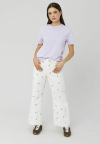 T-shirt lilas à manches courtes associé à un pantalon large blanc avec de petits motifs floraux. Des baskets sombres complètent la tenue.