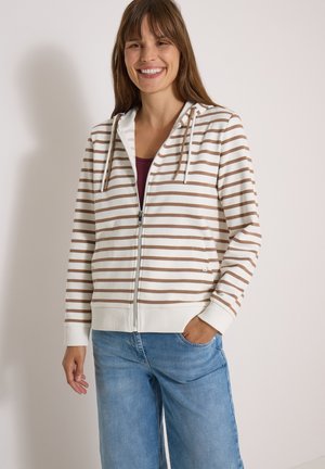 Femme souriante portant un sweat à capuche zippé blanc et marron à rayures avec cordons et un jean bleu, debout devant un fond clair uni.