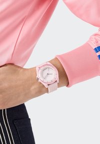 Roza ura z gladkim silikonskim trakom in okroglo številčnico, ki ima bele oznake in srebrne poudarke. Nenapetost na zapestju z roza rokavom.