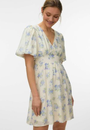 Kobieta w kremowej sukience o długości do kolan z niebieskim kwiatowym wzorem i bufkami, uśmiechająca się z zamkniętymi oczami na białym tle.