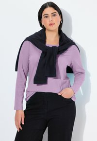 Helllila Langarmshirt, kombiniert mit schwarzen Hosen, dazu ein dunkler Pullover, der lässig um die Schultern gebunden ist. Glatter Stoff, entspannter Schnitt.