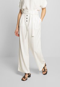 Vita randiga byxor med vida ben och hög midja, med bälte och svarta knappdetaljer på framsidan. Slät textur med lös passform.