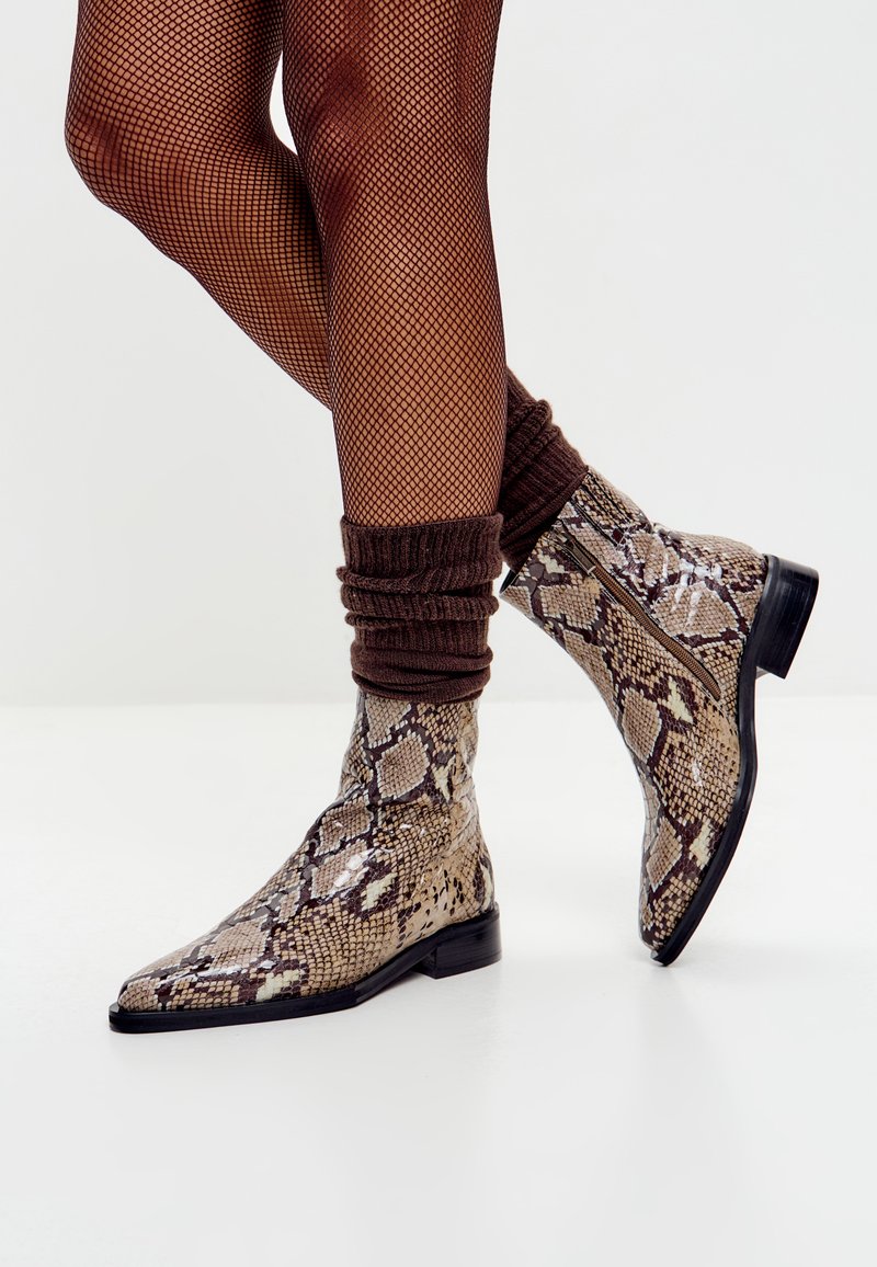 Botas de tobillo de piel de serpiente con punta afilada, que presentan tonos marrones y beiges, un acabado texturizado y una cremallera lateral. Acentuadas con calcetines marrones de canalé.