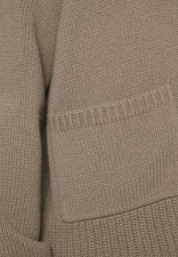 Beige gestrickter Pullover mit strukturiertem Finish, ausgestattet mit einer aufgesetzten Tasche und gerippten Details entlang der Kanten für zusätzliche Definition.