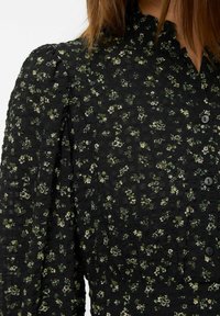 Zwart bloemenjurk met een getextureerde stof, groene en witte bloemenpatronen, een gerimpelde kraag en knoopsluitingen.