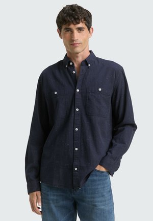Chemise boutonnée bleu foncé en tissu texturé, avec de légères speckles claires, deux poches poitrine et des boutons blancs, associée à un jean bleu.