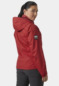 Vrouw met een zwarte paardenstaart, gekleed in een rode hoodiejack met het Helly Hansen-logo op de mouw en zwarte broek, kijkt over haar schouder.