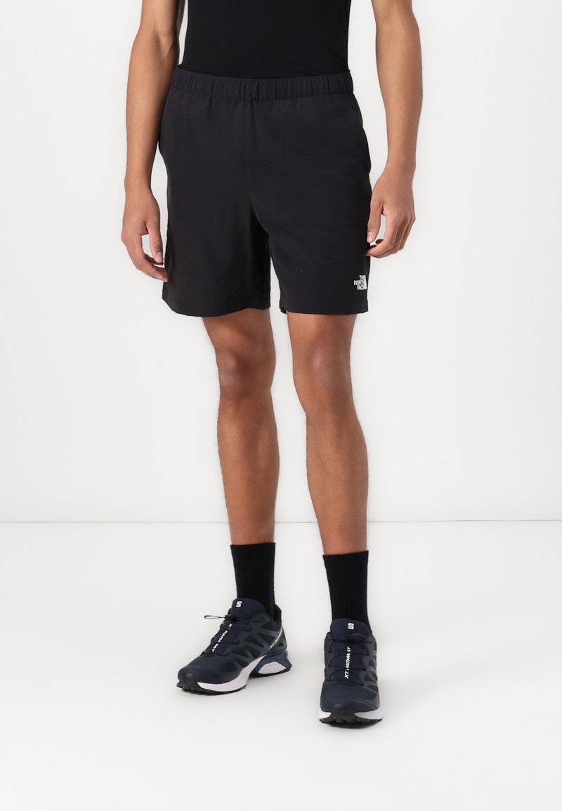 Sorte treningsshorts med elastisk liv, med en liten hvit logo. Brukt med sorte strømpesokker og mørke treningssko.