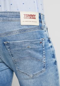 Vaaleansiniset farkut, joissa on takatasku, jossa on punavalkoinen "TOMMY JEANS" -merkki ja huomaamaton ompelu yksityiskohtia.