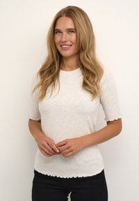 Vaaleanbeige lyhythihainen neulepaita, jossa on pyöreä kaula-aukko ja aaltoilevat reunat. Paita on pehmeästä materiaalista valmistettu ja muotonsa mukainen.