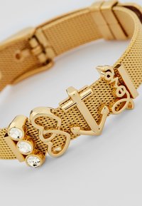 Bracciale in rete dorata con ciondoli, tra cui una croce e un'ancora, adornato con tre strass trasparenti. Design moderno con una finitura texturizzata.