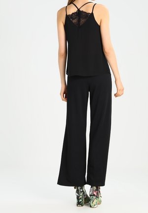 Kobieta ubrana w czarny, bezrękawnik z koronkowym tyłem, szerokie czarne spodnie i kwieciste botki na wysokim obcasie, widziana od tyłu.