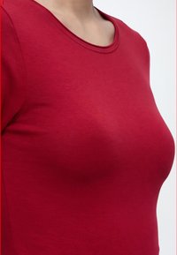 Gros plan d'une personne portant un t-shirt rouge uni à manches longues avec un col rond sur fond blanc.