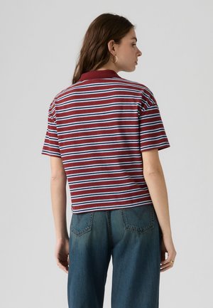 Femme aux longs cheveux bruns portant des boucles d'oreilles en créoles, un t-shirt rayé rouge et blanc à manches courtes et un jean bleu, de dos sur un fond uni.
