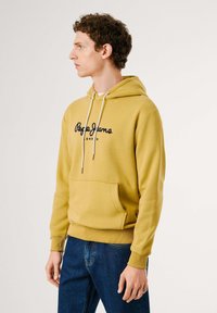 Yellow hooded sweatshirt with a front pocket, features black "Pepe Jeans London" logo, drawstring hood, and ribbed cuffs. Worn with blue jeans.