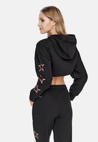 Felpa nera tagliata con dettagli a forma di stella rosa sulle maniche, abbinata a pantaloni jogger neri coordinati. Il materiale sembra morbido e testurizzato.