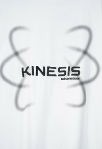 Biała tkanina z czarnym haftowanym napisem "KINESIS" oraz mniejszym napisem "podświadomość", otoczona delikatnymi szarymi abstrakcyjnymi pętlami po obu stronach.