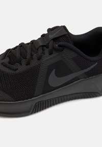 Zapatilla deportiva negra con parte superior de malla, paneles texturizados y suela de goma con surcos de tracción pronunciados. Logo de Nike en negro.