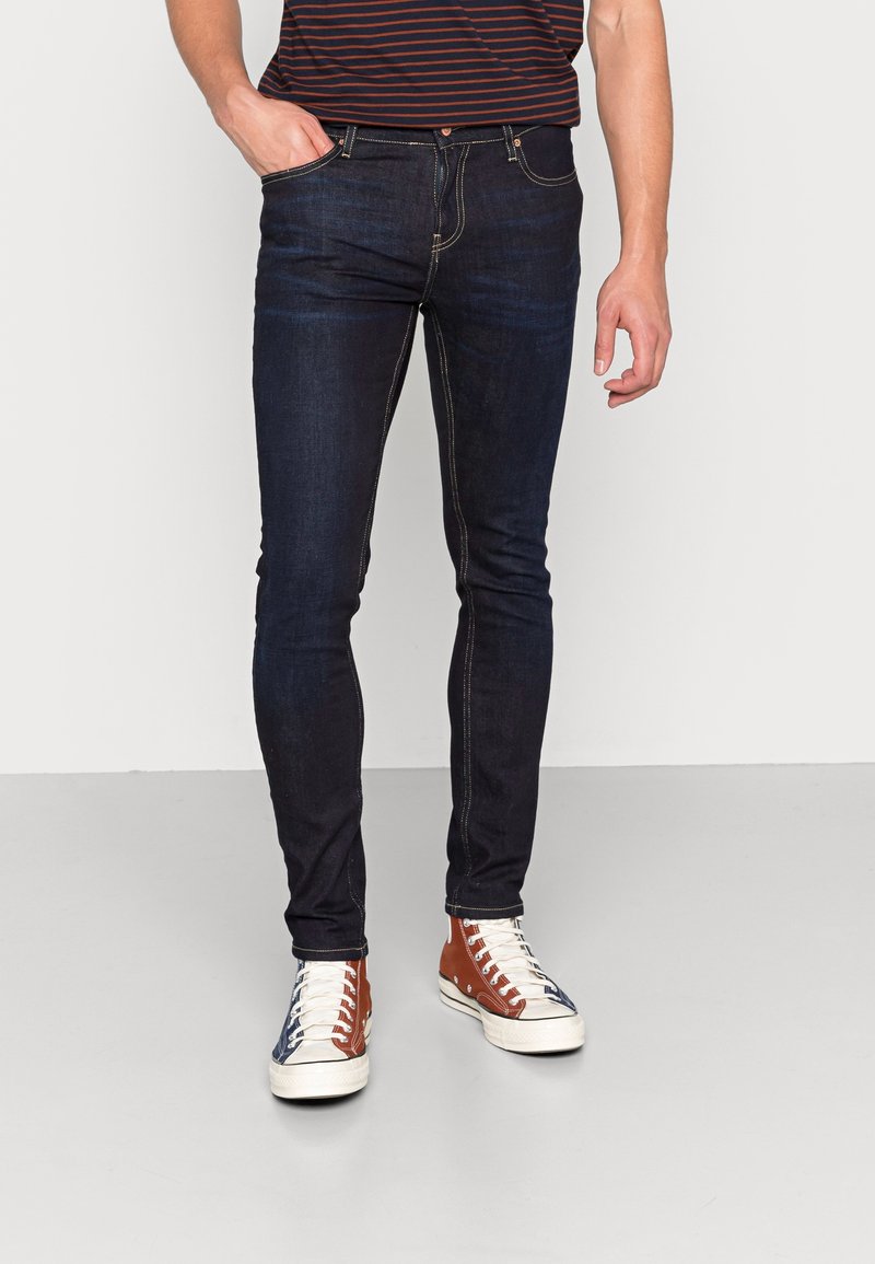 Mörkblå denimjeans med slim fit, med kontrasterande guldtråd, klassisk femfickdesign och bärs med bruna och vita sneakers.