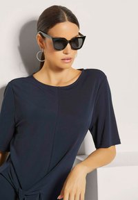 Navyblauwe jurk met korte mouwen, voorzien van een knoop in de taille en een zachte textuur. Zwarte hoekige zonnebril en ringoorbellen maken de look compleet.