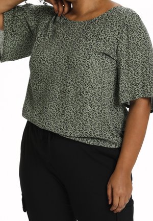 Personne portant un haut vert à manches courtes avec un petit imprimé floral noir et un pantalon noir, une main touche le cou et l'autre est le long du corps.